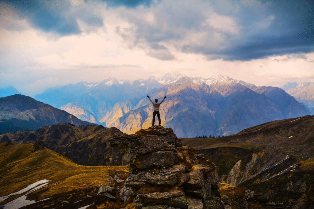 Man standing on a rocky mountain peak with arms raised overlooking a scenic valley. Republic Day long weekend