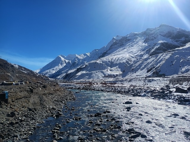 A river flowing through a rocky valley with snow-covered mountains under a bright sun.